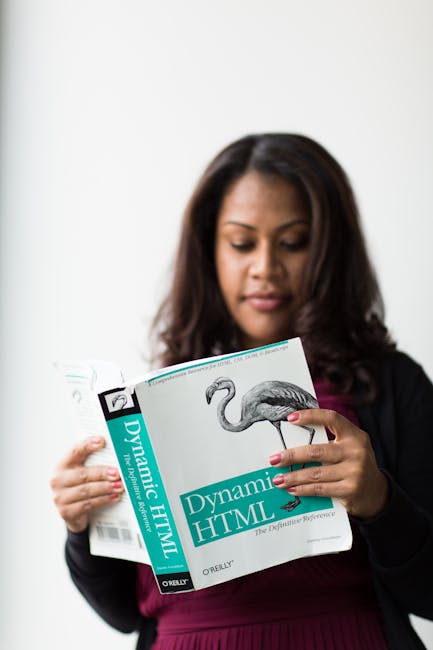 A woman with medium-length dark hair and light brown skin is holding a paperback book titled 'Dynamic HTML' with both hands. She is wearing a dark blazer over a purple dress and is standing in front o